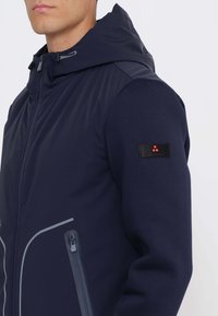 Uomo che indossa una giacca blu navy con cappuccio, con cerniera frontale e tasca con cerniera, piccola toppa nera con puntini rossi sulla parte superiore della manica.