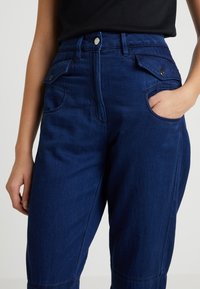 Navy blå högmidjade byxor i denim, med sidofickor, en framknapp och raka ben. Slät textur och rena linjer.