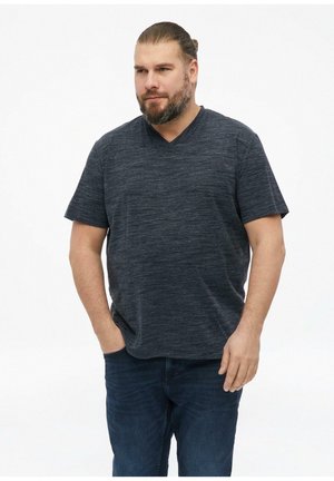 Brodaty mężczyzna z włosami związanymi do tyłu, ubrany w ciemnoszary t-shirt z dekoltem w serek i krótkim rękawem oraz niebieskie dżinsy, stojący z jedną ręką w kieszeni.
