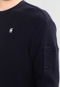 Nahaufnahme einer Person in einem dunkelblau strukturierten Langarmshirt mit einem kleinen weißen Logo auf der Brust und einer geknöpften Tasche am Ärmel.