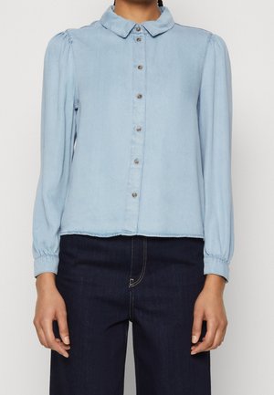 Jasnoniebieska koszula na długi rękaw z guzikami i marszczonymi mankietami, noszona z ciemnymi, wysokim stanem jeansami, na stojącej osobie.