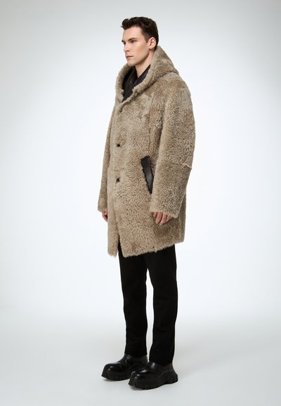 Beżowy płaszcz z kożuchem o futrzanej fakturze, z dużym kapturem i czarnymi skórzanymi akcentami. Noszony na czarnych spodniach i masywnych czarnych butach.
