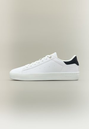 Sneakers basse bianche con lacci bianchi e dettaglio tallone nero, design minimalista, posate su uno sfondo beige neutro.