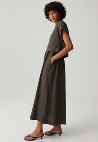 Woman with curly hair stands in profile wearing a dark green short-sleeve top and matching ankle-length skirt with pockets and black heeled sandals.