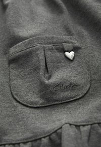 Szary materiał o teksturowanej fakturze, z kieszenią i zawieszką w kształcie serca. Na dolnej części wyszyte jest "Clarks".