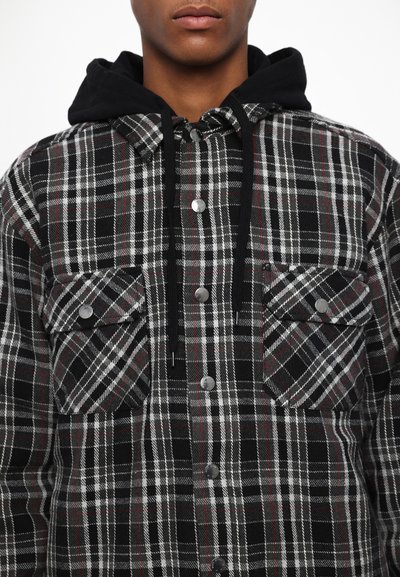 Veste en flanelle à carreaux noirs et gris avec des accents rouges, boutons-pression et capuche noire. Comprend deux poches poitrine à rabat.
