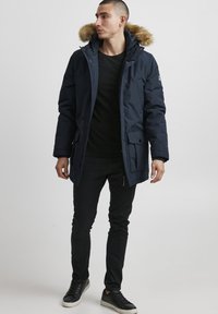 Mørkeblå parkas med pelsforet hette, med frontlommer og glidelås. Bæret over en svart skjorte, kombinert med mørke bukser og joggesko.