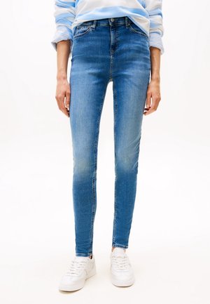 Person iført blå skinny jeans, hvide sneakers og en blå og hvid stribet skjorte med opkrogede ærmer, stående mod en ensfarvet hvid baggrund.