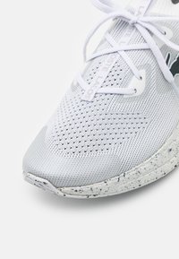 Baskets de sport blanches avec empeigne en mesh, détails texturés, bout arrondi et semelle intermédiaire gris moucheté. Lacets et accents du logo en gris clair.