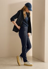 Navy athletic pants with zippered pockets, paired with a blue sweatshirt and sleeveless black vest, topped with a blue cap and white sneakers.