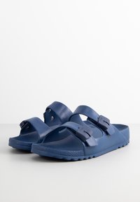 Blue plastic sandals with two adjustable straps and circular accents. Textured sole for grip, lightweight design, and an open-toe style.