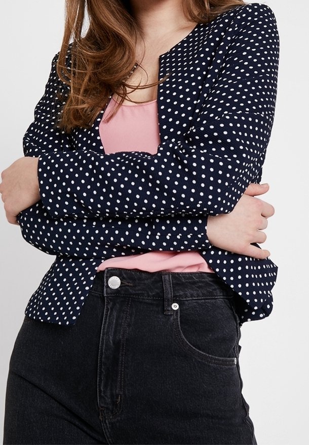 Blazer bleu marine à pois avec une coupe ajustée, haut rose clair en dessous, et jean en denim noir. Caractéristiques : pois blancs, manches longues et encolure.
