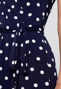 Granatowa sukienka kopertowa w białe groszki, o gładkiej fakturze i zawiązanym paskiem w talii.