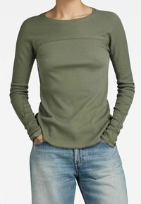 Olivgrön långärmad topp med ribbat mönster, rund halsringning och figurnära passform. Bärs tillsammans med ljusblå jeans, med synliga söm detaljer.