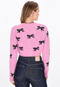 Suéter corto rosa hecho de un material suave, con patrones de lazo negro. Tiene mangas largas y un cuello redondo, combinado con jeans de denim oscuro.
