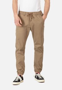 Pantalones jogger color tan con cintura elástica y cordón. Presentan puños elásticos y bolsillos laterales. Combinados con zapatillas negras y blancas.