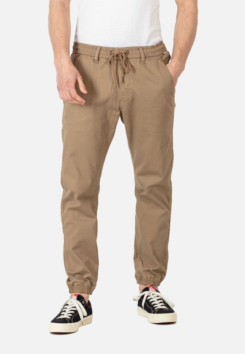 Pantalones jogger color tan con cintura elástica y cordón. Presentan puños elásticos y bolsillos laterales. Combinados con zapatillas negras y blancas.