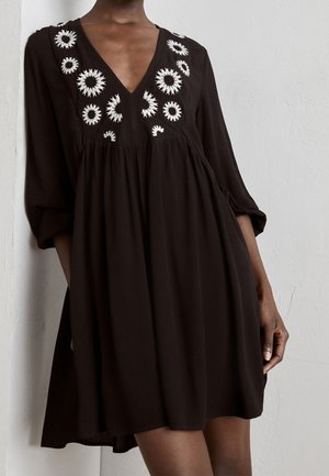 Czarna sukienka z długim rękawem, głębokim dekoltem w kształcie litery V i białymi, okrągłymi haftowanymi wzorami wokół dekoltu i górnej części klatki piersiowej.