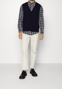 Navy knit vest over a checked shirt, paired with white trousers and black lace-up shoes. Features a v-neck and button detailing.