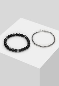 Bracciale nero con perline e accenti in argento accanto a un bracciale a catena in argento. Il bracciale con perline presenta texture lisce opache, mentre la catena ha maglie intrecciate.
