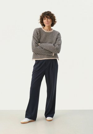 Pull en chevrons tricoté beige et bleu marine, accompagné d'un pantalon large bleu marine. Des baskets blanches avec des semelles texturées complètent la tenue.