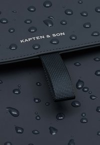 Zwarte waterdichte tas met verhoogde waterdruppels, voorzien van een gestructureerde schouderband en een zilveren logo van "KAPTEN & SON" op de voorkant.
