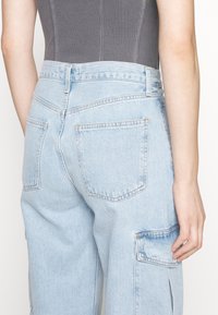 Osoba oblečená v jasně modrých džínech s zadními kapsami a kapsou na boční straně, v kombinaci se šedým žebrovaným topem.