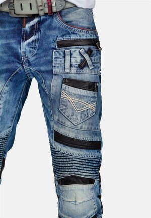 Denim cargo pants in light blue. Features multiple pockets, black leather accents, and stitched patterns. Textured detailing along the legs.