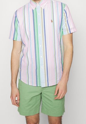 Homme portant une chemise à manches courtes à rayures pastel verticales et un short vert, debout devant un fond blanc.