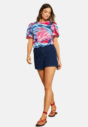Kobieta ubrana w bluzkę z bufkami o abstrakcyjnym wzorze w kolorach czerwonym, niebieskim i różowym, ciemnoniebieskie szorty oraz czerwone, płaskie sandały na paskach, stojąca na białym tle.