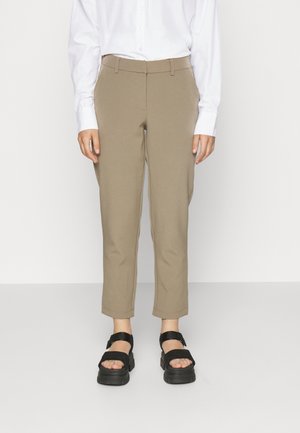 Person iført beige cropped bukser, hvid langærmet skjorte og sorte platformsandaler stående mod en ensfarvet baggrund.