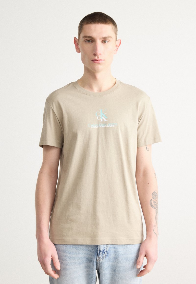 Camiseta de algodón beige con cuello redondo que presenta un texto de logo frontal con "Calvin Klein Jeans" en azul claro. Manga corta y un corte clásico.