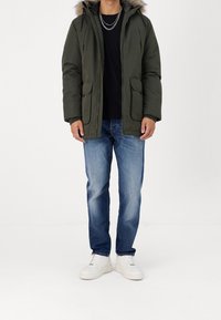 Grön vadderad parkas med pälskantad huva, två framfickor och dragkedja, kombinerad med blå jeans och vita sneakers.