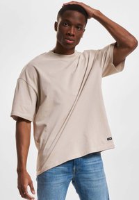 T-shirt oversized beige in morbido tessuto, con maniche corte e scollatura a girocollo. Dettaglio etichetta all'orlo. Indossato con jeans blu.