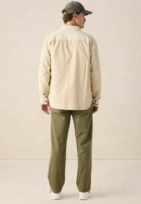 Beige overhemd met lange mouwen en knoopmanchetten, gecombineerd met olijfgroene broek, op een persoon die tegen een neutrale achtergrond staat.