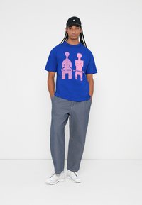 Blå t-skjorte med rosa grafisk design som viser to figurer og teksten "de nye originalene," sammen med grå vide bukser og hvite joggesko.