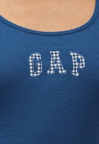 Μπλε ριμπ αμάνικη μπλούζα με τα γράμματα «GAP» σε λευκό καρό μοτίβο στο στήθος.