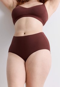 Kurvige Person, die braune hochgeschnittene Unterwäsche und einen passenden BH trägt, steht vor einer schlichten, hellen Wand.