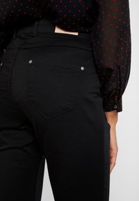 Černé džínové kalhoty s přiléhavým střihem, se jednou zadní kapsou ve tvaru špičky a zlatými kovovými akcenty.