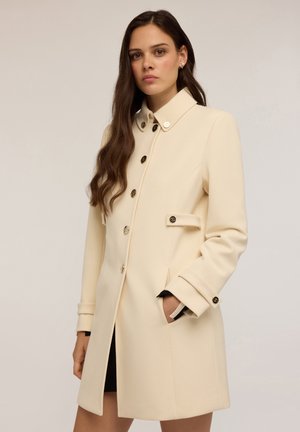 Trench-coat crème avec un design structuré, présentant des boutons dorés, des poches latérales et un col. Tissu lisse et texturé.
