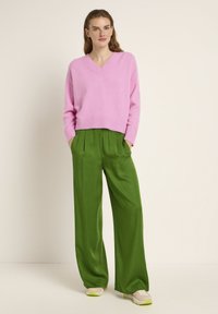 Rosa Cashmere-V-Ausschnittpullover mit geripptem Saum, kombiniert mit grünen Satin-Hosen mit weitem Bein. Das Model trägt leichte Sneakers mit grünen Akzenten.