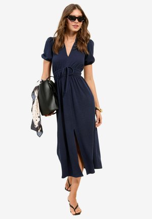 Kobieta ubrana w granatową sukienkę midi z dekoltem w serek i bufkami na rękawach, czarne klapki, okulary przeciwsłoneczne, niesie czarną torebkę z apaszką w groszki.