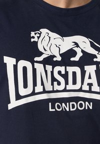 Tricou din bumbac bleumarin, cu un grafic mare al unui leu alb și textul îndrăzneț "LONSDALE LONDON", având un guler clasic la gât și o textură moale.
