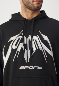 Felpa nera con cappuccio caratterizzata da un prominente logo iridescente "Jordan" e il testo "Sport". Il design include una classica tasca a marsupio e corde di serraggio.