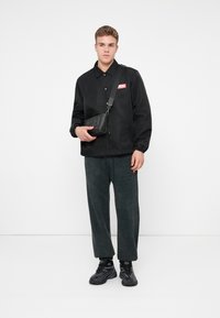 Junger Mann, der eine schwarze Knopfjacke, graue lockere Hosen, schwarze Sneakers und eine schwarze Umhängetasche in einem weißen Studio trägt.