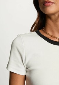 Maglietta bianca a coste con scollo rotondo nero e maniche corte. Inquadratura ravvicinata che evidenzia la texture e il contrasto di colore con una collana a catena.