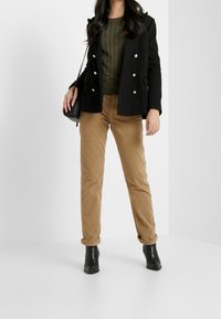 Schwarzer zweireihiger Mantel mit silbernen Knöpfen, grüner Kabelstrickpullover, beige Cordhosen und schwarze Stiefeletten. Durchgehend glatte Texturen.