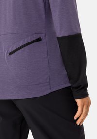 Maglia lunga viola con dettagli neri, dotata di tasca con zip e tessuto testurizzato, abbinata a pantaloni neri eleganti.