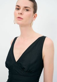 Sort kjole med dyb V-udskæring, tekstureret stof, tætsiddende silhuet og subtil samling ved taljen. Model iført markante øreringe.