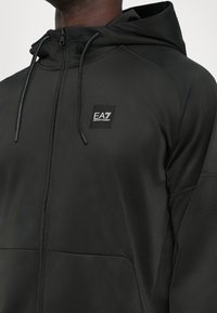 Svart zip-up hoodie i mjukt tyg, med en framsidan logotypapplikation, justerbar dragsko huva och sidofickor.